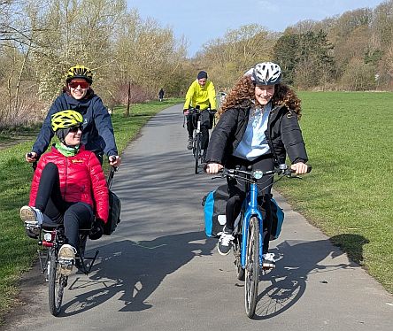Vier Personen fahren auf einem Radweg durch eine Wiesenlandschaft, Bäume und Hügel im Hintergrund