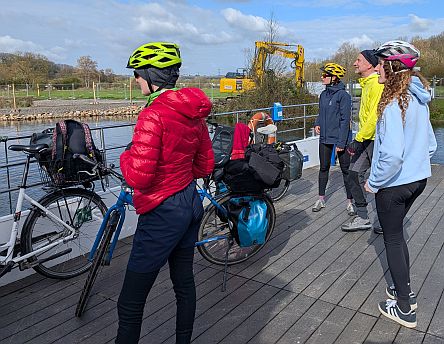 Vier Personen mit Fahrradhelmen stehen auf einer kleinen Fähre, im Hintergrund ein Fluss und Bäume