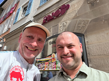Zwei Personen machen ein Selfie vor einem Imbissladen mit Speisekarten an der Wand