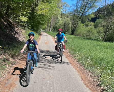 Zwei Kinder fahren mit Fahrrädern auf einem schmalen asphaltierten Weg durch ein bewaldetes Tal