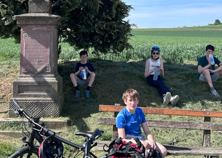 Mehrere Kinder sitzen im Schatten eines Denkmals an einer Bank und machen eine Pause, Fahrräder liegen davor