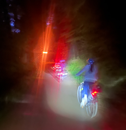 Person fährt nachts mit beleuchtetem Fahrrad auf einem dunklen Waldweg, Rücklicht leuchtet rot
