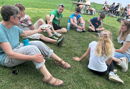 Gruppe von Erwachsenen und Kindern sitzt auf einer Wiese im Kreis, einige essen von Tellern