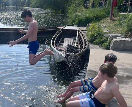 Mehrere Kinder springen und sitzen an einem Seeufer, ein Ruderboot liegt am Steg