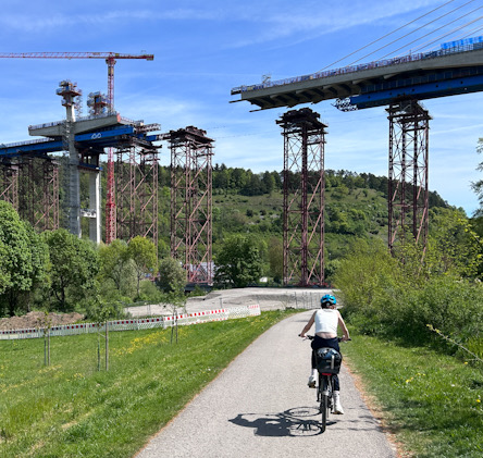 Person fährt mit Reiserad auf einem Weg unter einer im Bau befindlichen Brücke mit hohen Stahlpfeilern