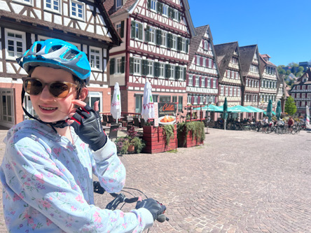 Kind mit Fahrradhelm und Sonnenbrille sitzt auf einem Fahrrad auf einem historischen Marktplatz mit Fachwerkhäusern