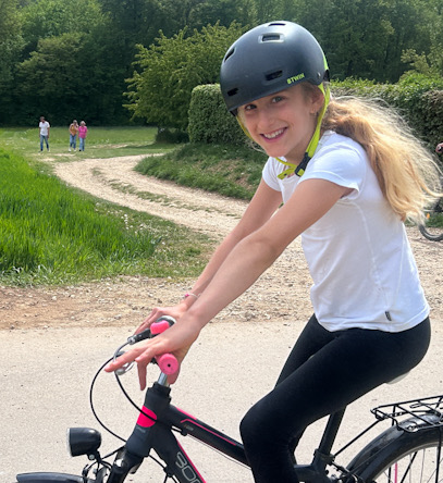 Kind mit Fahrradhelm lächelt auf einem Fahrrad an einem Feldweg, weitere Personen im Hintergrund