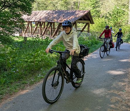 Mehrere Kinder fahren mit Fahrrädern auf einem Weg durch einen Wald, eine überdachte Holzbrücke im Hintergrund