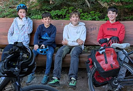 Vier Kinder sitzen auf einer Holzbank im Wald, Fahrräder und Helme liegen davor