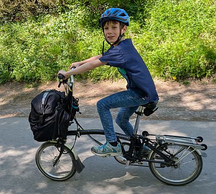 Kind fährt ein kompaktes Reiserad mit Gepäck auf einem asphaltierten Weg durch grünes Umfeld