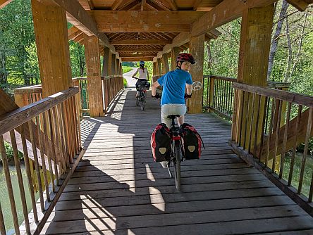 Mehrere Personen fahren mit Fahrrädern über eine überdachte hölzerne Brücke im Wald