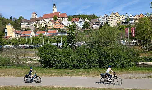 Zwei Personen fahren mit Fahrrädern auf einem Weg entlang eines Flusses, Stadt mit Kirche im Hintergrund