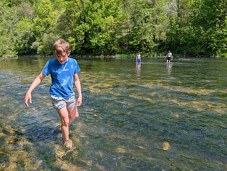 Kind watet barfuß durch einen flachen Fluss mit klarem Wasser, weitere Personen im Hintergrund