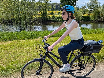 Person fährt mit bepacktem Fahrrad auf einem Weg entlang eines Flusses mit grüner Ufervegetation