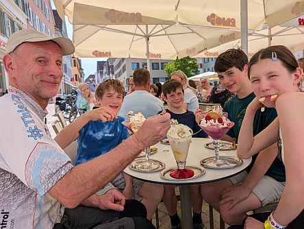 Gruppe von Personen sitzt an einem runden Tisch im Außenbereich eines Cafés und isst Eisbecher