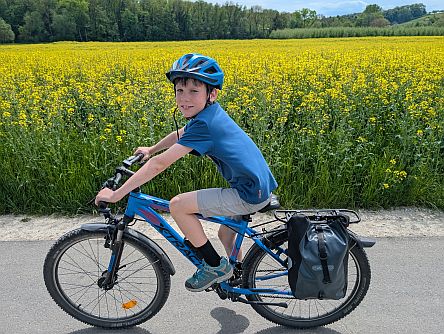 Kind fährt mit bepacktem Fahrrad auf einem Asphaltweg entlang eines gelb blühenden Rapsfeldes