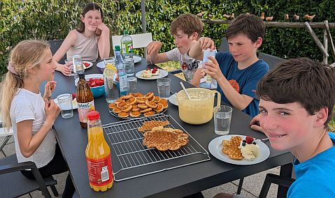 Mehrere Kinder und eine erwachsene Person sitzen an einem Gartentisch mit Waffeln, Obst und Getränken